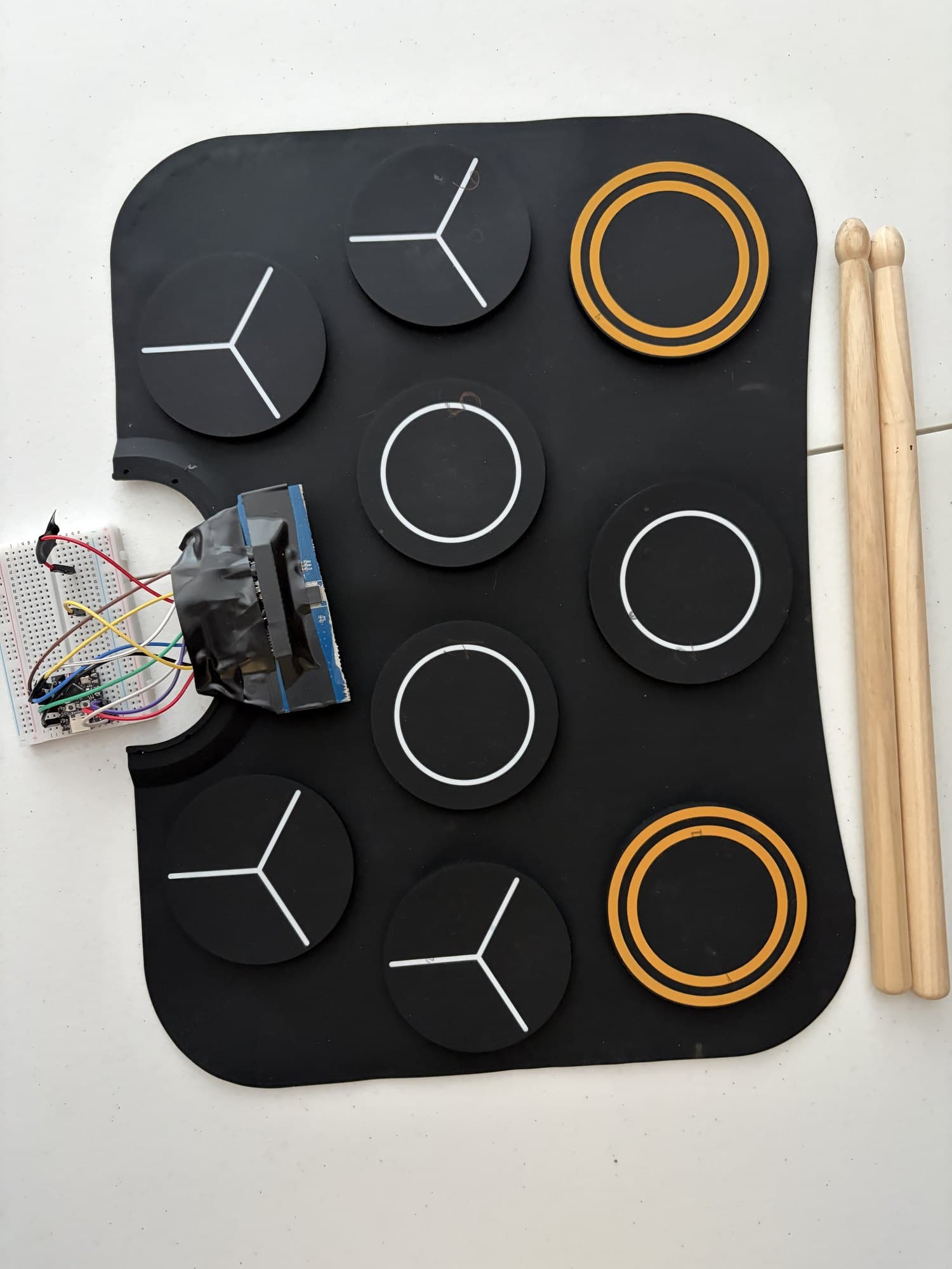 Drum Cockpit from above: a black nine-pad drum mat with white and orange rings, a taped ESP32 board wired to a breadboard on the left, and two drumsticks.