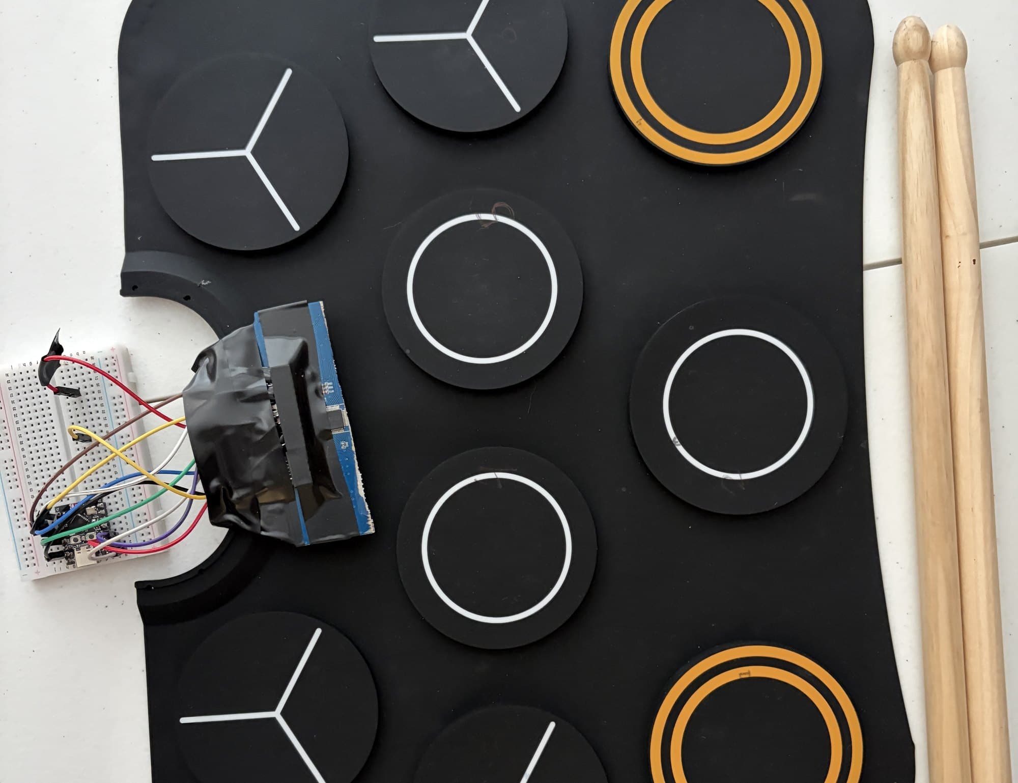 Close-up of the drum mat's edge: colored jumper wires running from the pads' taped connector to an ESP32-C3 board on a white breadboard, with drumsticks beside the mat.