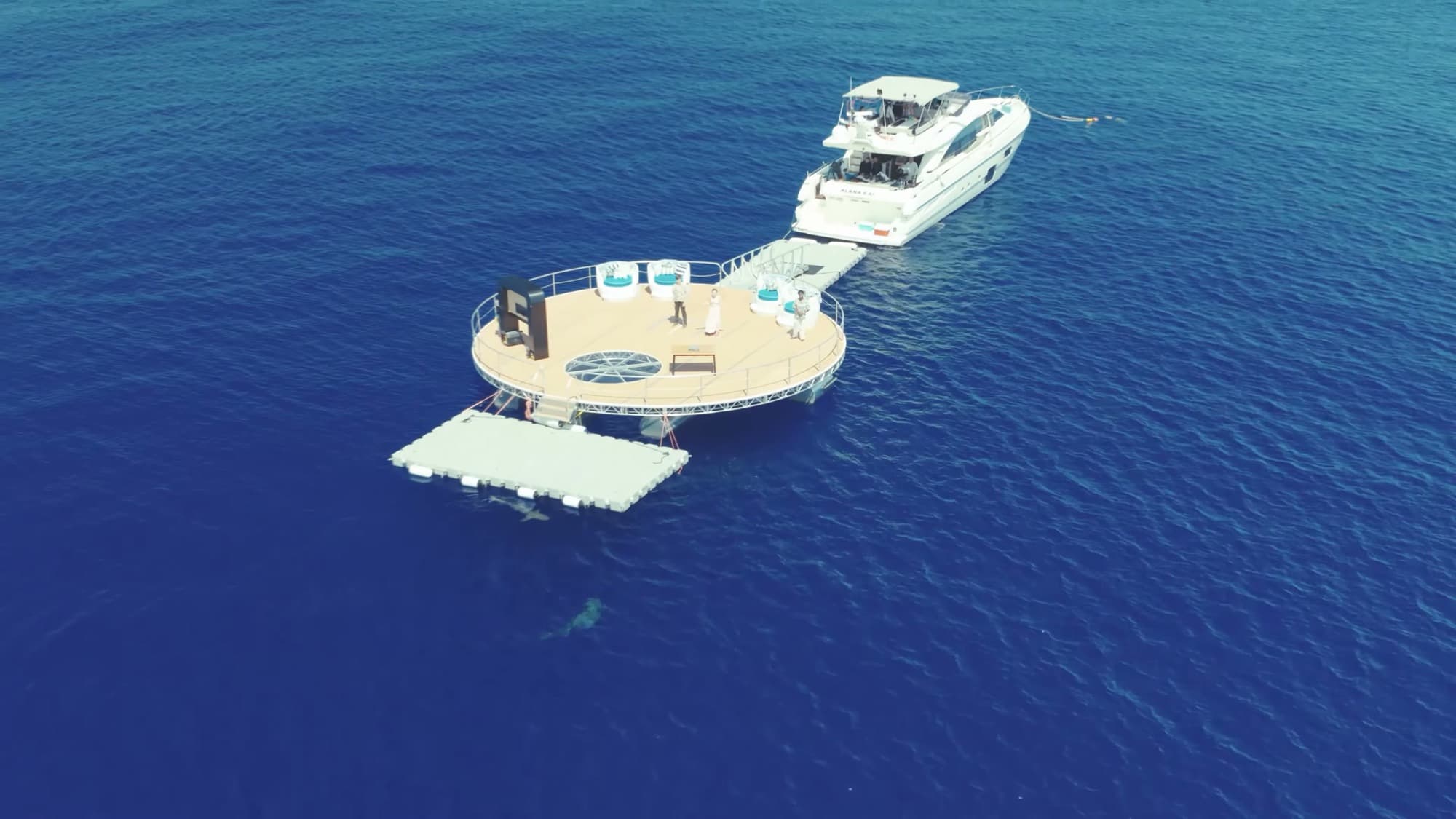 Aerial view of the round pitch platform and a yacht moored together in deep blue water.