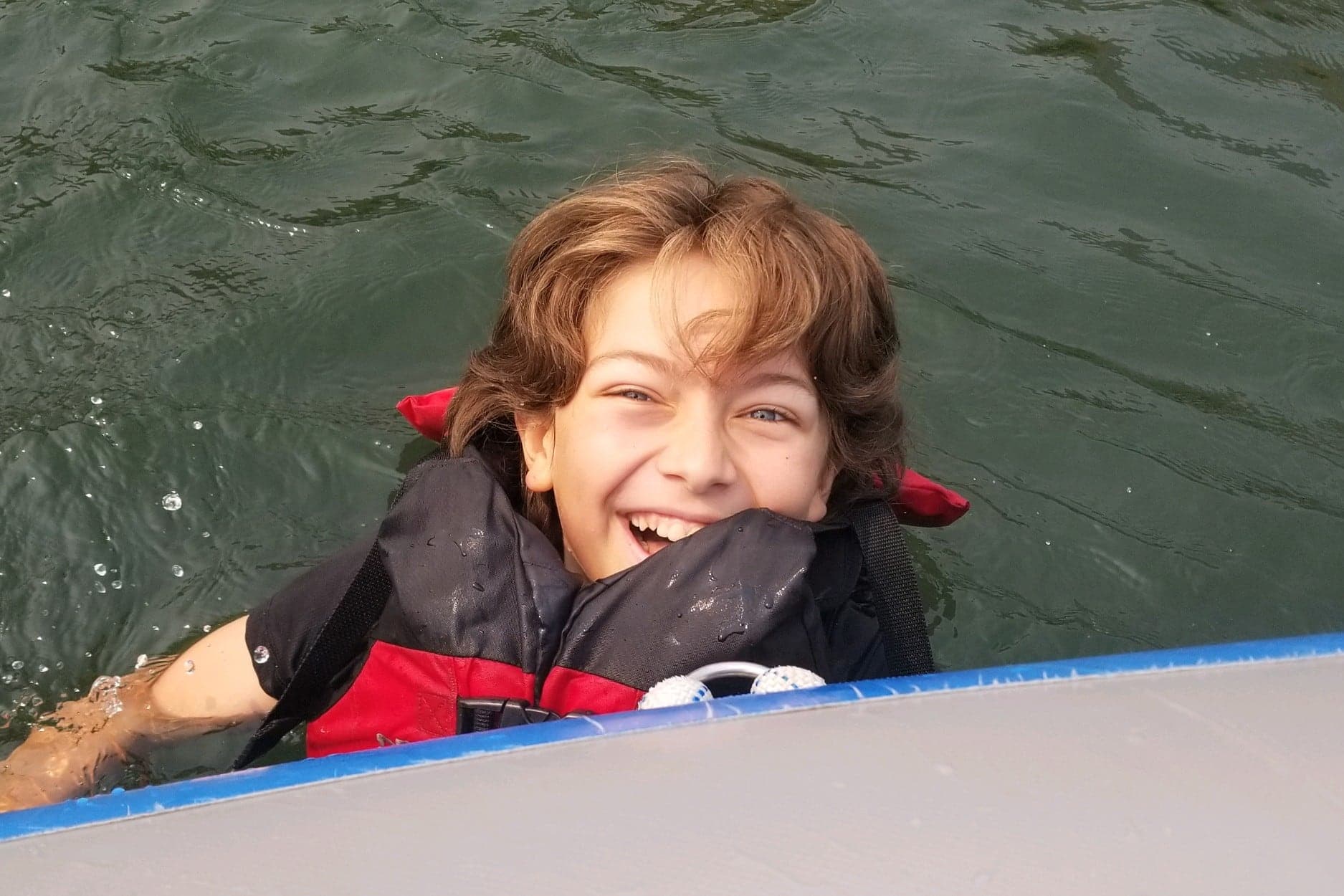 Cruce at 12, laughing in a lake in a red and black life jacket, holding on to the edge of an inflatable float.